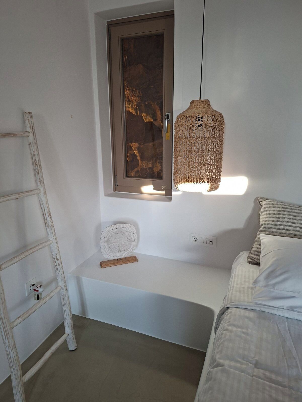 A bedroom with a white bed, a wooden ladder, a wall-mounted shelf with a decorative plate, and a woven light fixture.