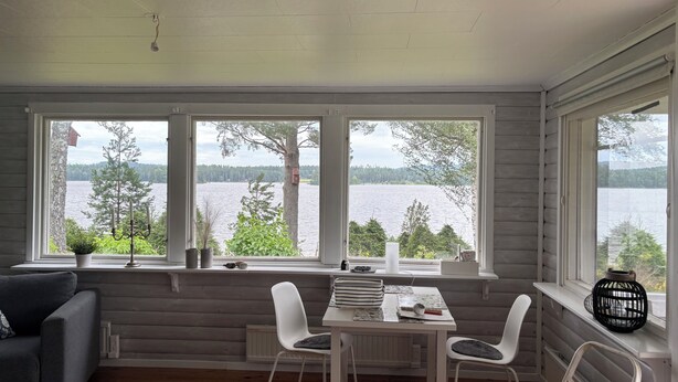 A room with a table and chairs, a window with a view of a lake and trees, and a grey couch. - Swedish house directly on the lake with motorboat & own boat ramp (Storfors)
