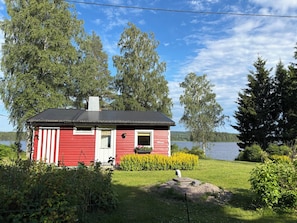 Eine rote Holzhütte mit schwarzem Dach, umgeben von viel Grün und einem See.