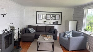 A modern living room with a grey sofa, a grey armchair, a coffee table, and a fireplace. There are framed pictures on the wall and a window with a view of greenery. - Swedish house directly on the lake with motorboat & own boat ramp (Storfors)