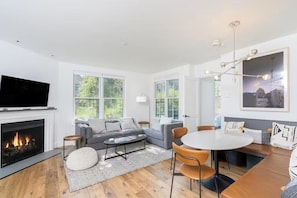 A modern living room with a fireplace, grey sofa, round dining table, and large windows.