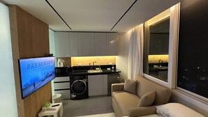 Ett modernt kök med en inbyggd ugn, en diskho och en kaffemaskin. På väggen sitter en tv som visar programmet ”PASSENGER home”. En soffa står placerad nära ett fönster med utsikt över staden på natten.