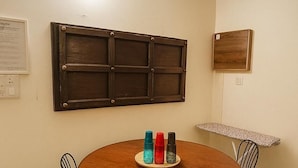 Une table de salle à manger ronde en bois, accompagnée de quatre chaises, un luminaire fixé au mur, un tableau d’affichage et une petite poubelle.