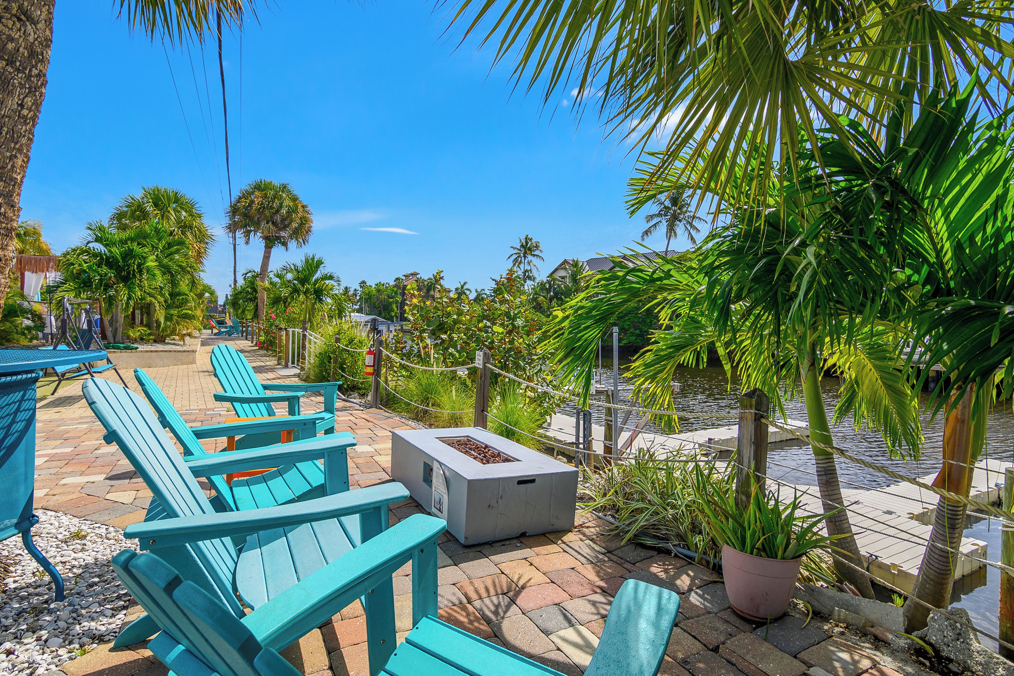 Outdoor seating with chairs and a fire pit, surrounded by palm trees and a water body.