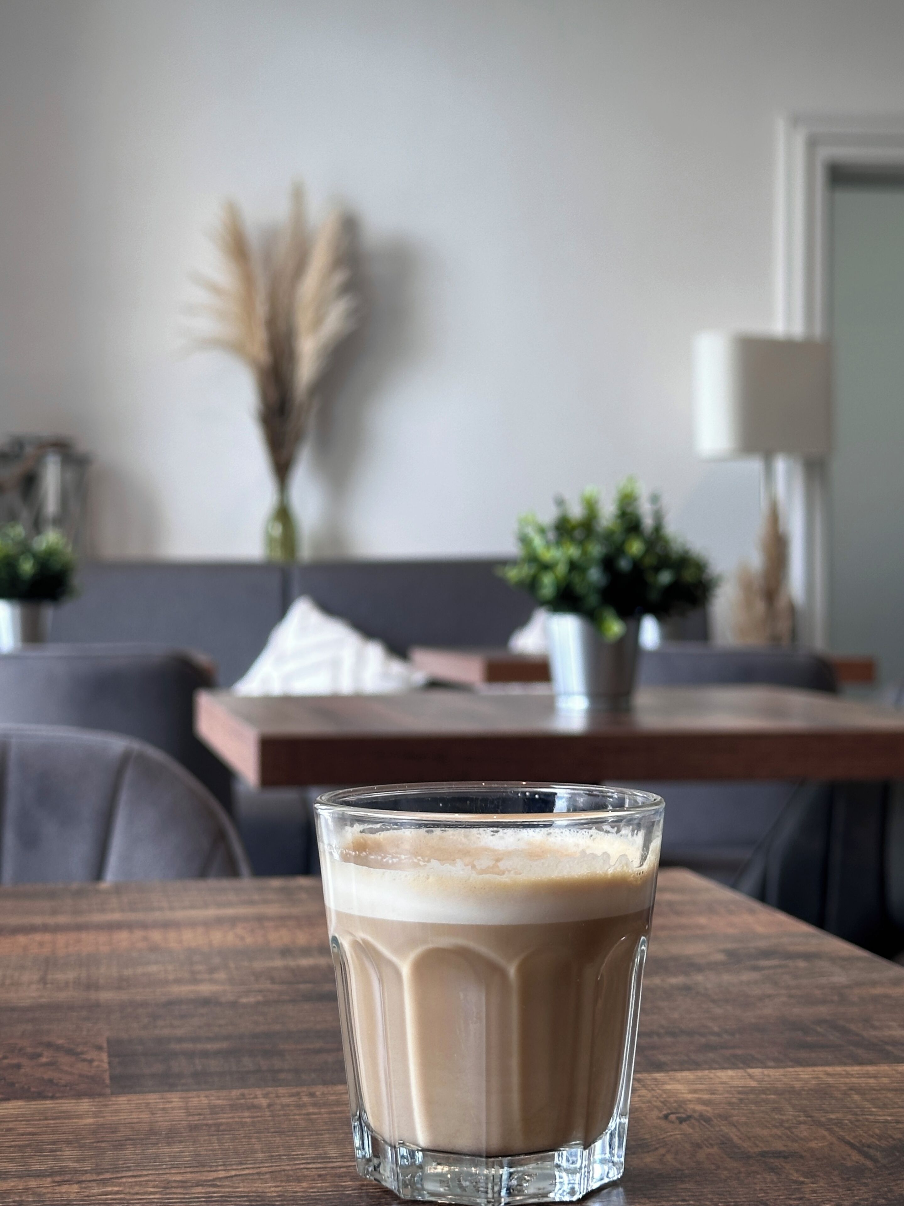 Uma chávena de café transparente sobre uma mesa de madeira numa sala de estar moderna, com um sofá, uma lâmpada e plantas decorativas.