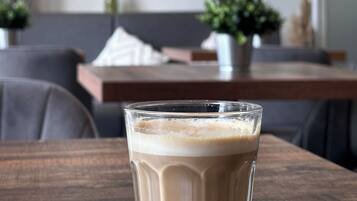 Uma chávena de café transparente sobre uma mesa de madeira numa sala de estar moderna, com um sofá, uma lâmpada e plantas decorativas.