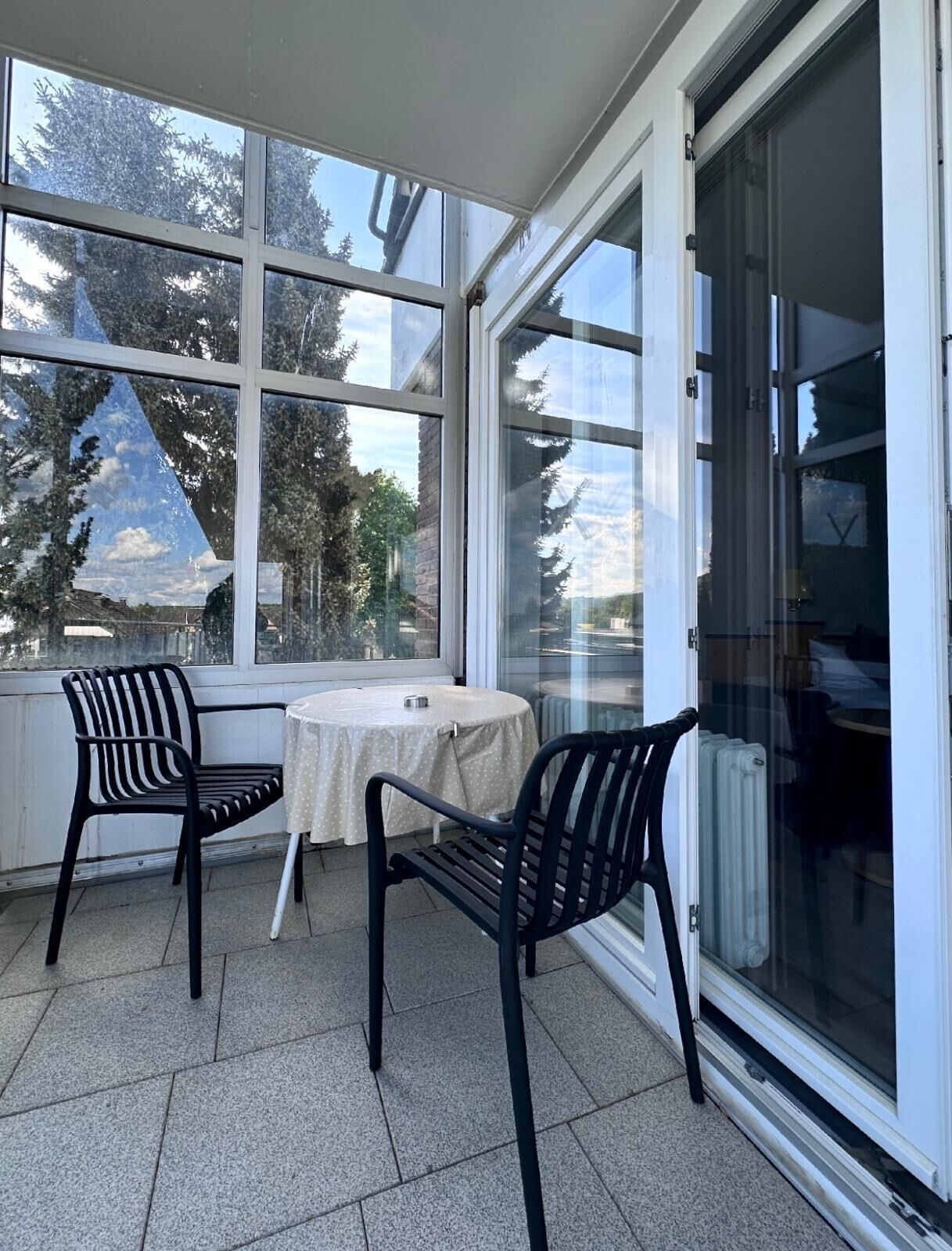 Une terrasse couverte avec deux chaises et une table, de grandes fenêtres et une vue sur des arbres et un bâtiment.