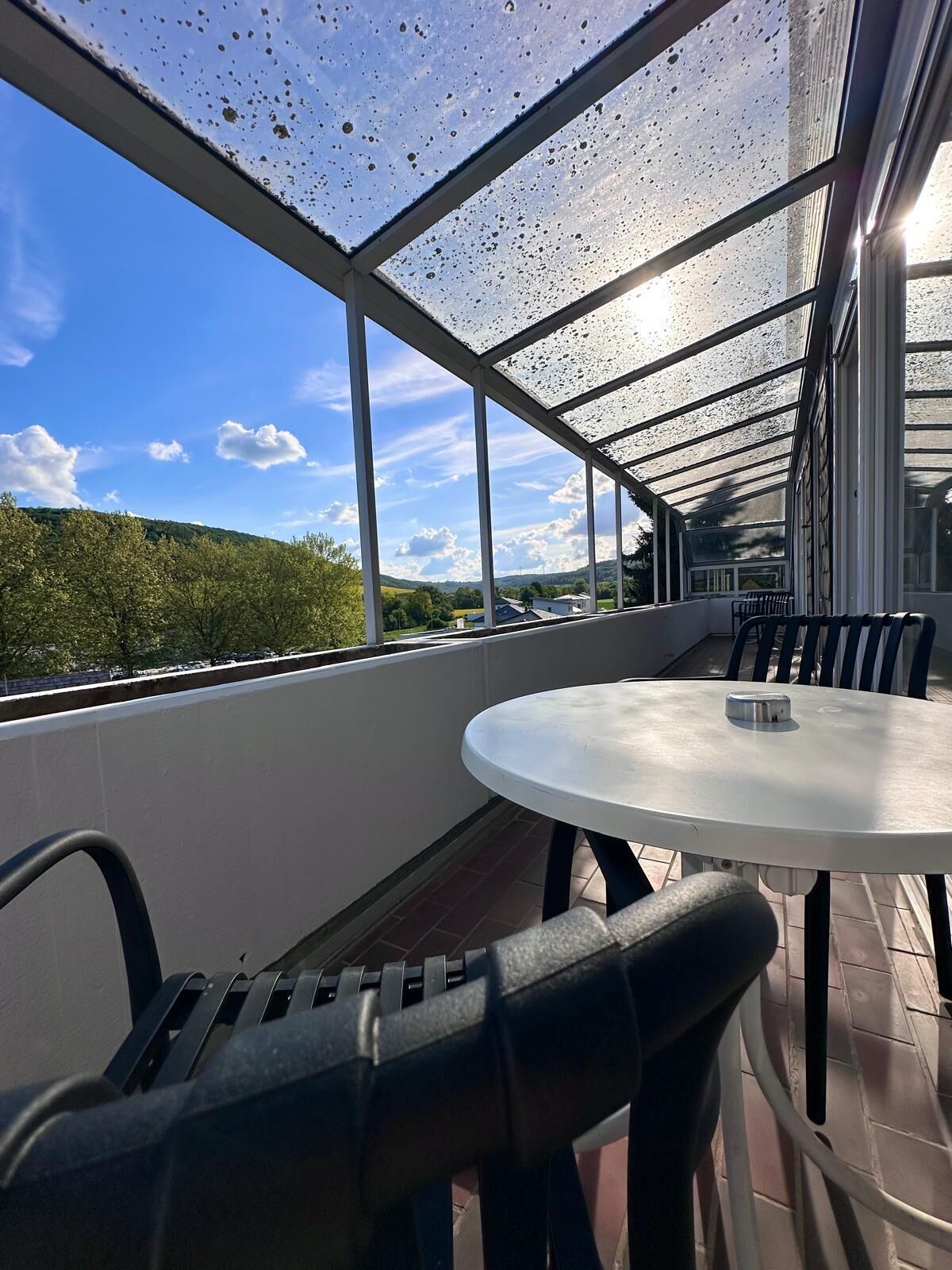 A covered outdoor seating area with a table and chairs, large windows, and a view of greenery and hills.