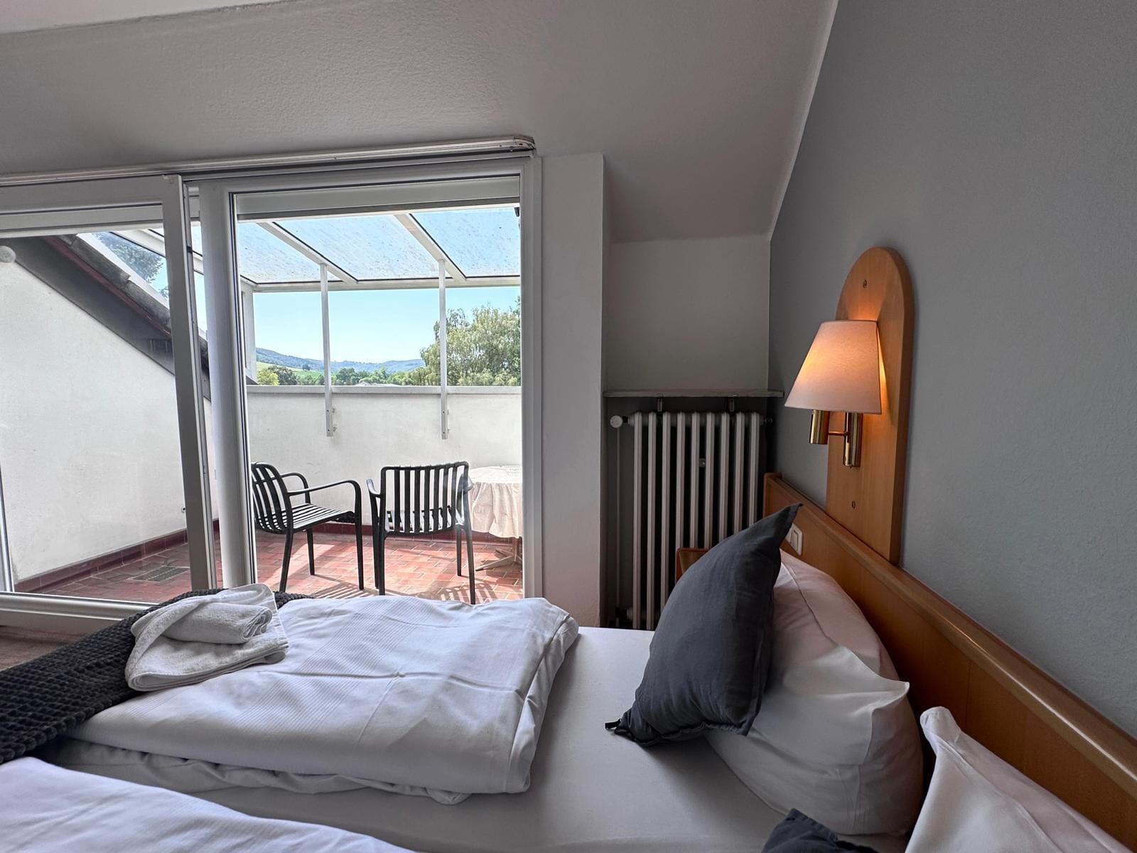 Een hotelkamer met een bed, nachtkastje, lamp, raam en uitzicht op de omgeving.