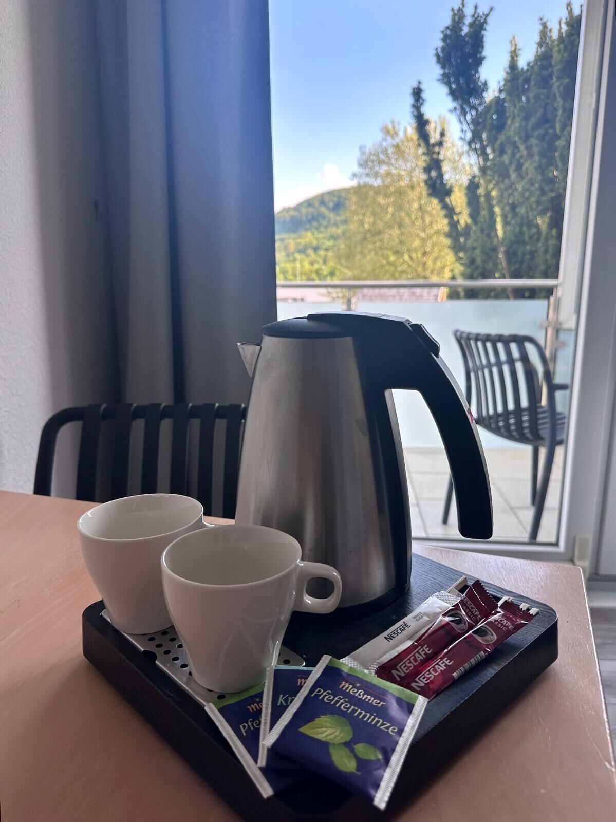 Een dienblad met twee witte bekers, een roestvrijstalen koffiepot en pakjes Nescafe koffie op een tafel bij een raam met uitzicht op bomen en een balkon.