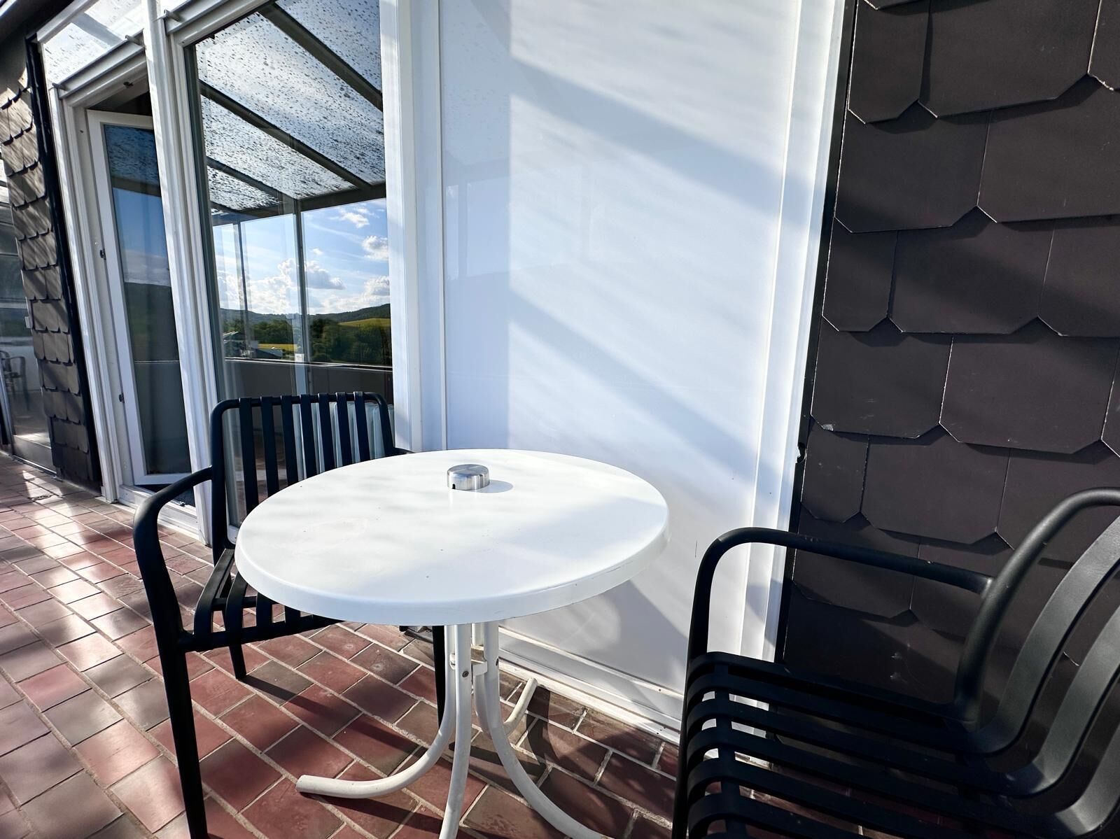 Une terrasse avec une table ronde et deux chaises, une porte vitrée donnant sur l’extérieur et un sol carrelé.