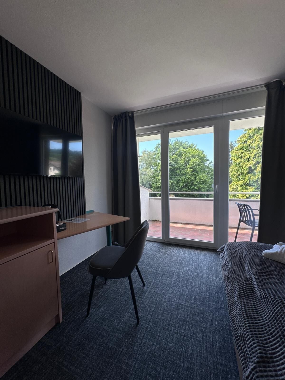 Een hotelkamer met een bureau, stoel, bed, televisie en een balkon met uitzicht op bomen.