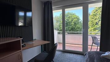 Een hotelkamer met een bureau, stoel, bed, televisie en een balkon met uitzicht op bomen.