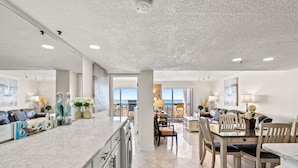 A spacious kitchen with a marble countertop, dining area with wooden chairs, and a view of the ocean through large windows. - Pristine Direct Oceanfront Getaway Luxury Oasis (North Myrtle Beach)
