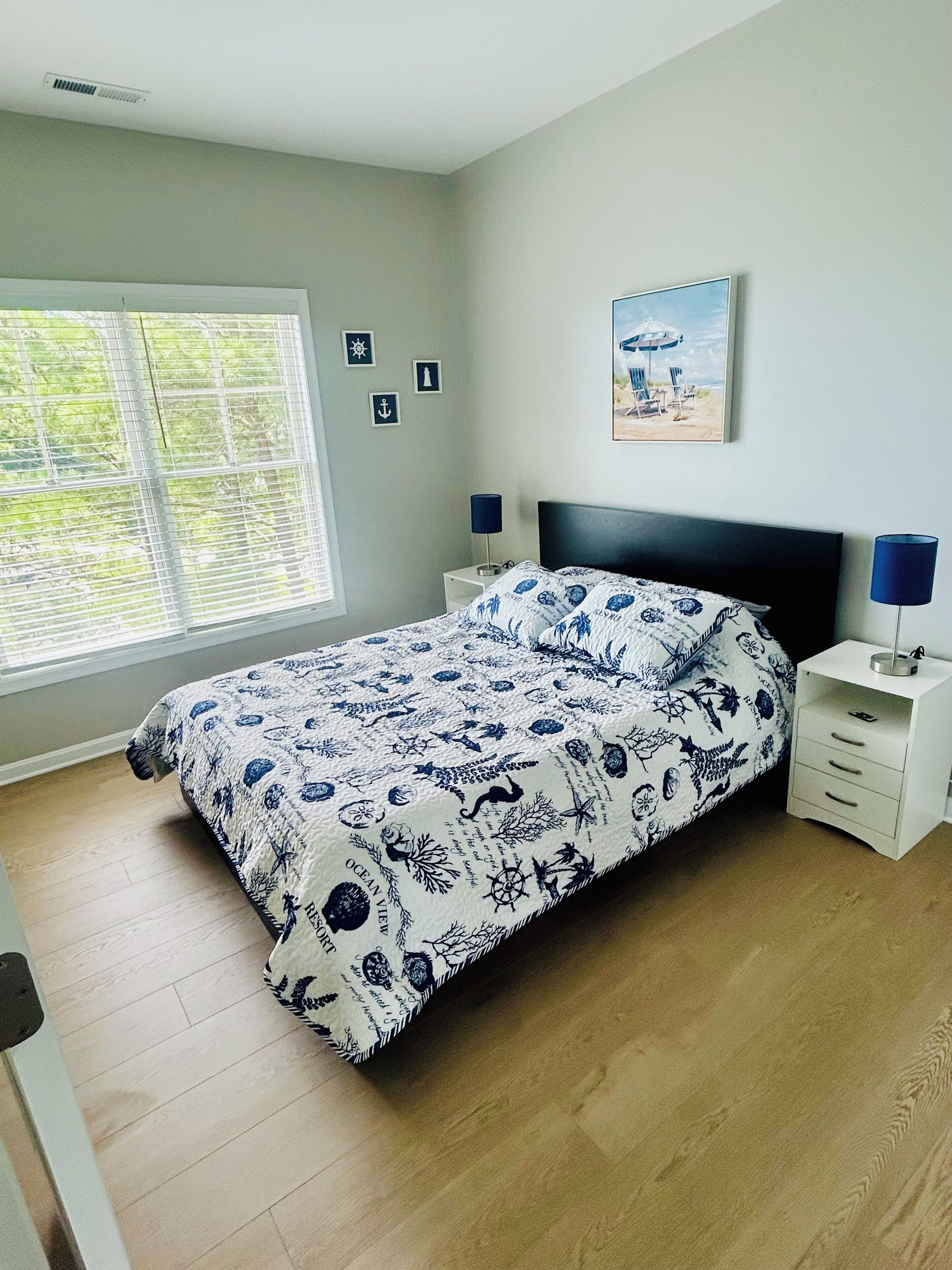 A bedroom with a bed, two bedside tables, a window with blinds, and a wall-mounted picture.