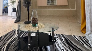 Een moderne woonkamer met een salontafel met glazen blad, een vloerventilator, een flatscreen-televisie aan de muur en een klein schapje met decoratieve objecten.