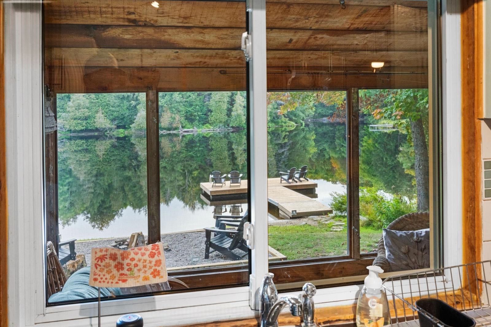 A cabin with a large window overlooking a lake and dock, wooden interior, and a view of the surrounding forest.