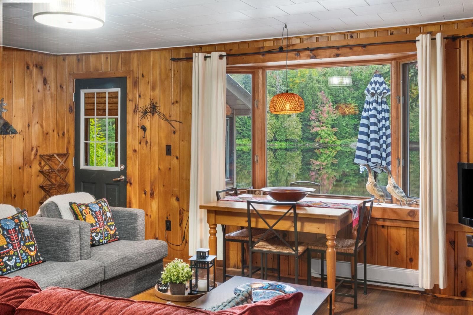 A cozy cabin interior with a dining area, wooden walls, a sofa with colorful pillows, and a view of trees through the window.