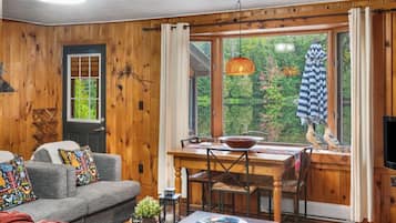 A cozy cabin interior with a dining area, wooden walls, a sofa with colorful pillows, and a view of trees through the window.