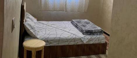 Un dormitorio pequeño con cama individual, un taburete de madera y una ventana con cortinas de encaje.