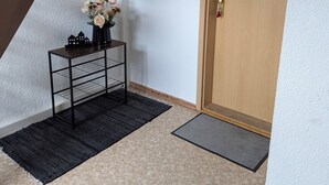 Un couloir avec une porte en bois, une console en métal noir sur laquelle est posé un vase de fleurs, un tapis noir et un petit paillasson.