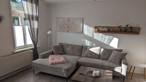Un salon moderne avec un canapé d’angle gris, une petite table basse ronde et une étagère en bois agrémentée d’objets décoratifs.
