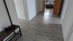 Un couloir avec un banc en bois et une pancarte de bienvenue. Il y a une porte sur la gauche et, au fond, une pièce avec un lit et une fenêtre.