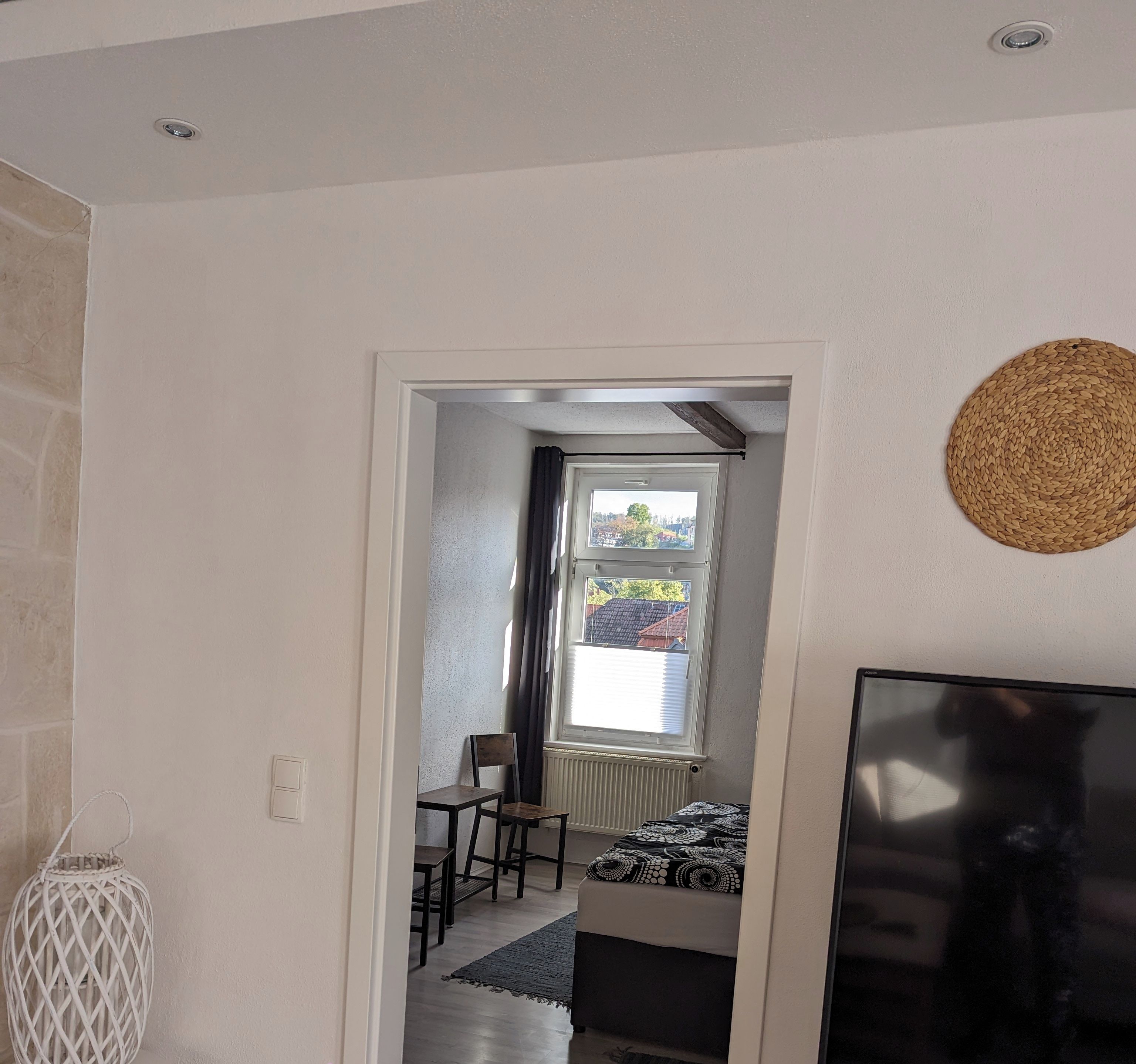 Un salon moderne avec un téléviseur, une tapisserie murale tissée et une vue sur un espace salle à manger, accessible par une porte ouverte.
