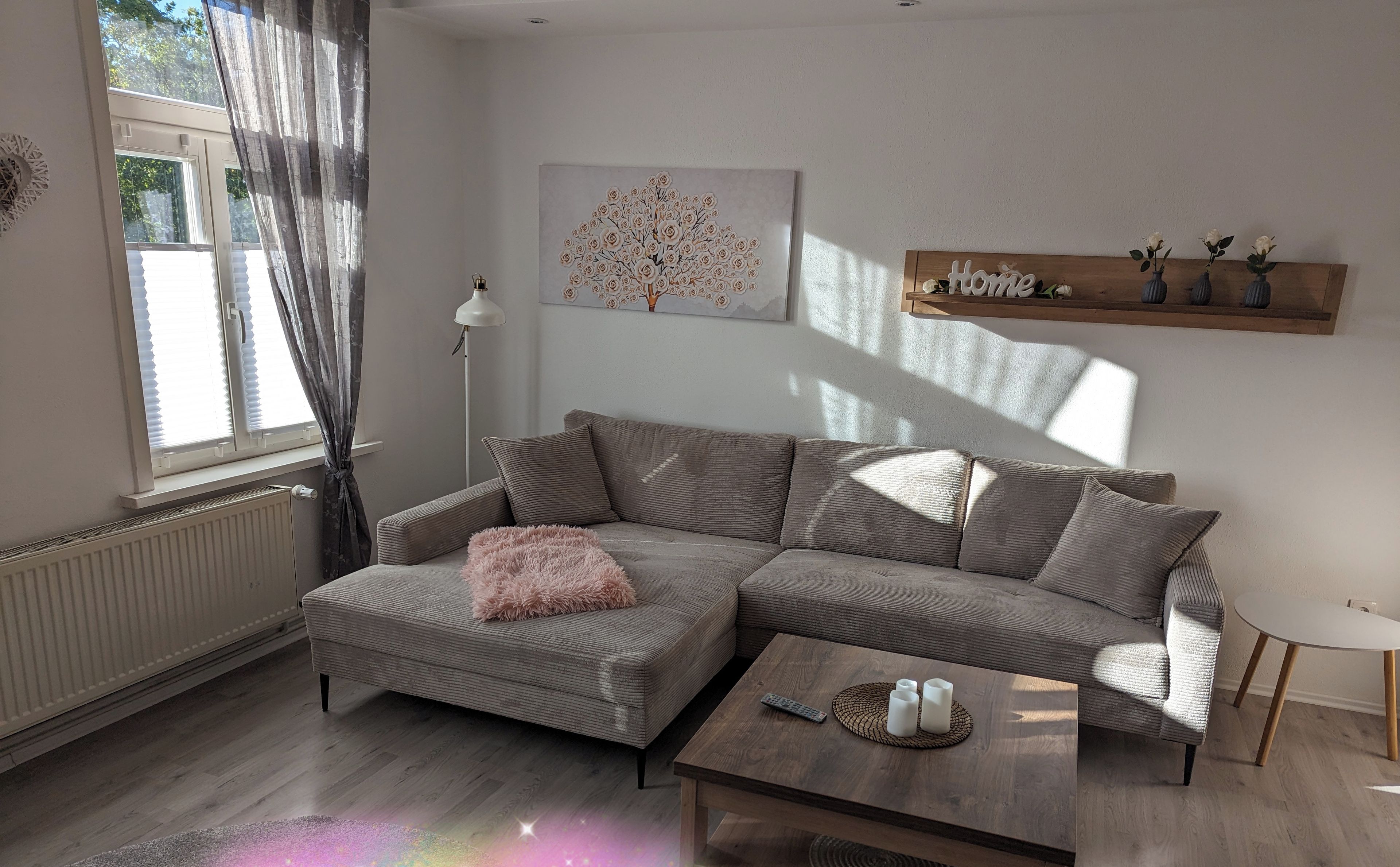 Een moderne woonkamer met een grijze hoekbank, een klein rond salontafeltje en een houten plank met decoratieve objecten.