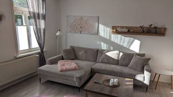 Een moderne woonkamer met een grijze hoekbank, een klein rond salontafeltje en een houten plank met decoratieve objecten.