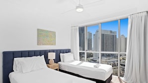 A hotel room with two beds, a large window with a city view, a ceiling fan, and a decorative wall piece. - 12th Floor Ocean Views in Chevron Towers (Surfers Paradise)
