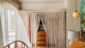 Un dormitorio con cama, una silla de madera, cortina de macramé, una lámpara y vista al exterior.