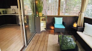 Un cuarto acogedor con una puerta corrediza de vidrio que da a una cocina, un sofá de mimbre con cojines, una mesita y vistas al bosque a través de amplias ventanas.