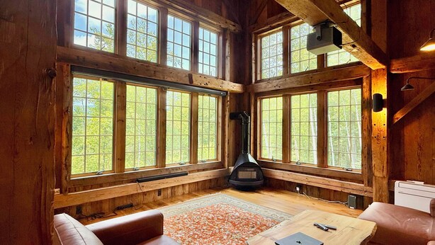 Interior rústico de una cabaña con ventanas amplias, una chimenea, sofás de cuero y una mesa de madera.