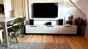 A modern living room with a flat-screen TV mounted on the wall, a white entertainment unit with speakers, a dining table with chairs, and a decorative vase with dried plants. - Cozy Flat in Villa Near Old Town (București)