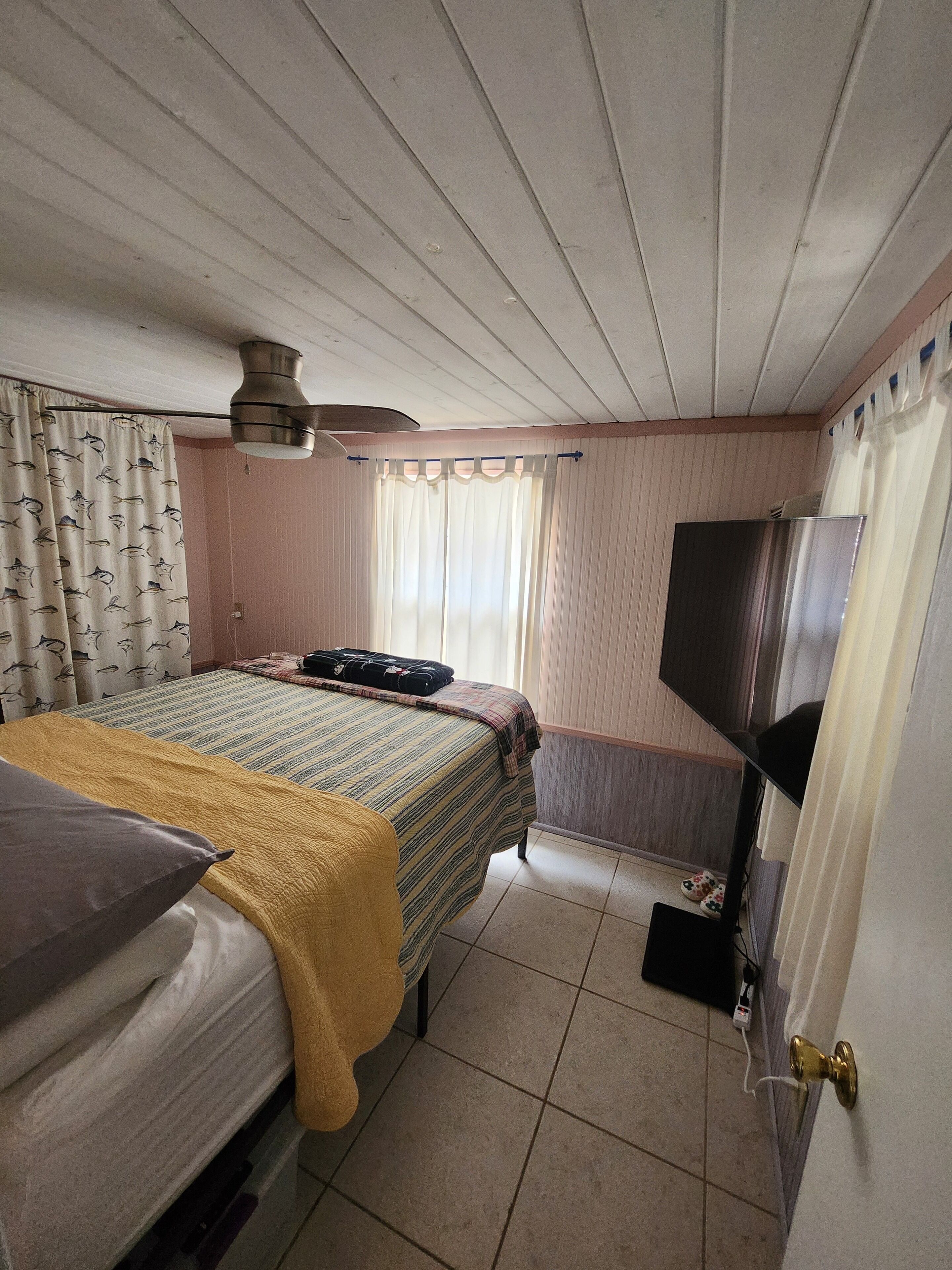 A bedroom with a bed, a television, a window with curtains, and a ceiling fan.