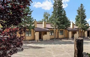 A row of single-story buildings with a flat roof, surrounded by trees and a gravel ground.