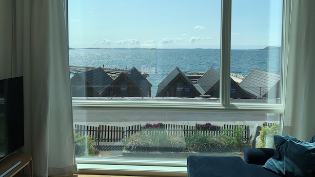 Ein Zimmer mit großem Fenster mit Blick auf eine Küstenlandschaft, Holzbauten und einem klaren Himmel.