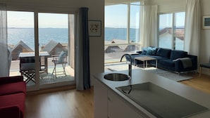 Ein modernes Wohnzimmer mit einer großen Schiebeglastür, die auf einen Balkon mit Blick auf das Meer und Strandhäuser führt. Es gibt ein blaues Sofa, einen kleinen Tisch und eine Küchenzeile mit Spüle.