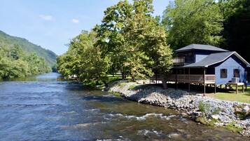 Een vakantiehuis aan de rivier met een terras, omringd door bomen en een rotsachtige oever.