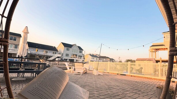A person reading a book in a wicker chair with a view of a beach and houses in the background. - Unique Home on Peaceful Waterfront (Stafford Township)
