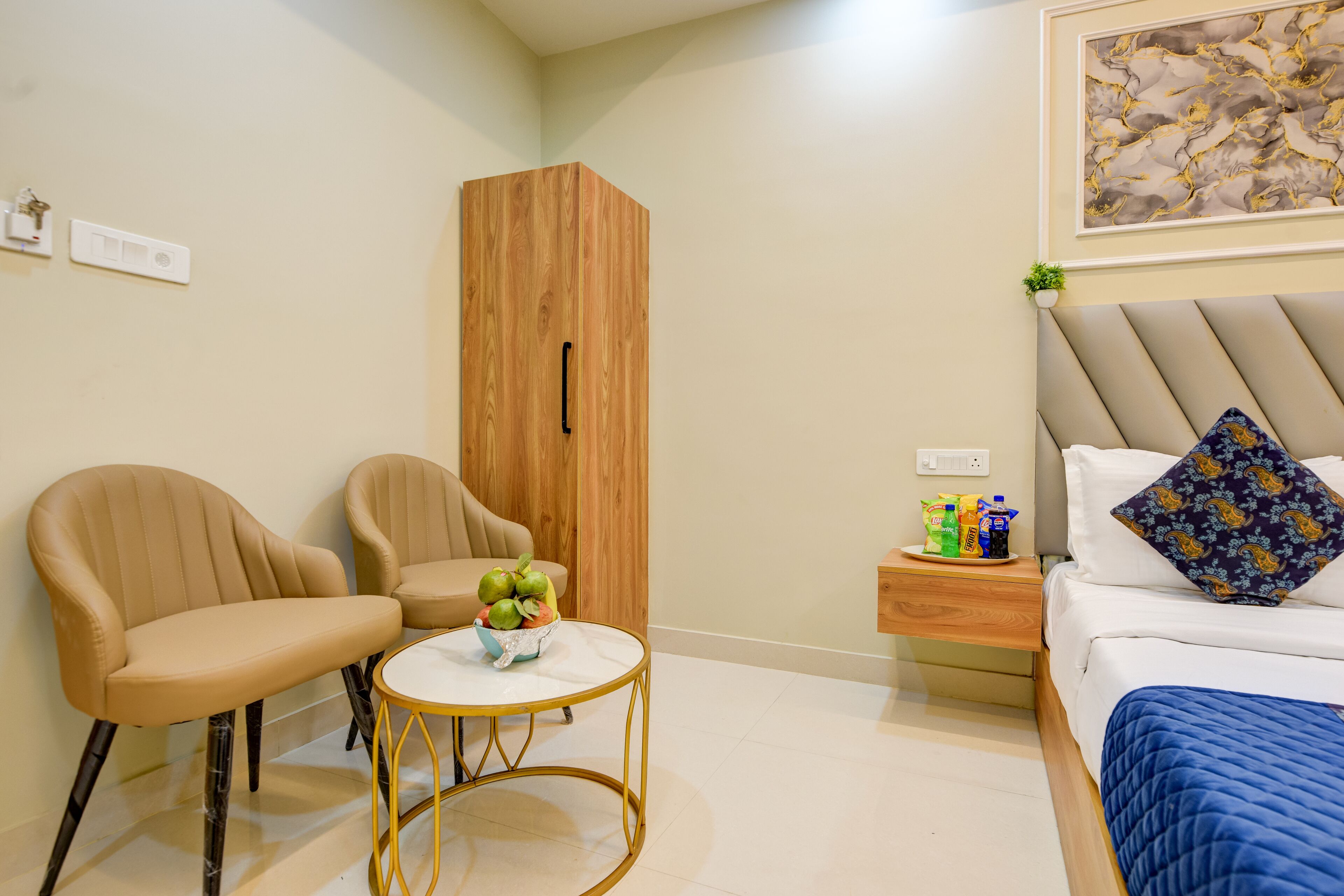 A modern hotel room with a bed, two chairs, a small table with fruit, a nightstand with snacks, and a wooden wardrobe.