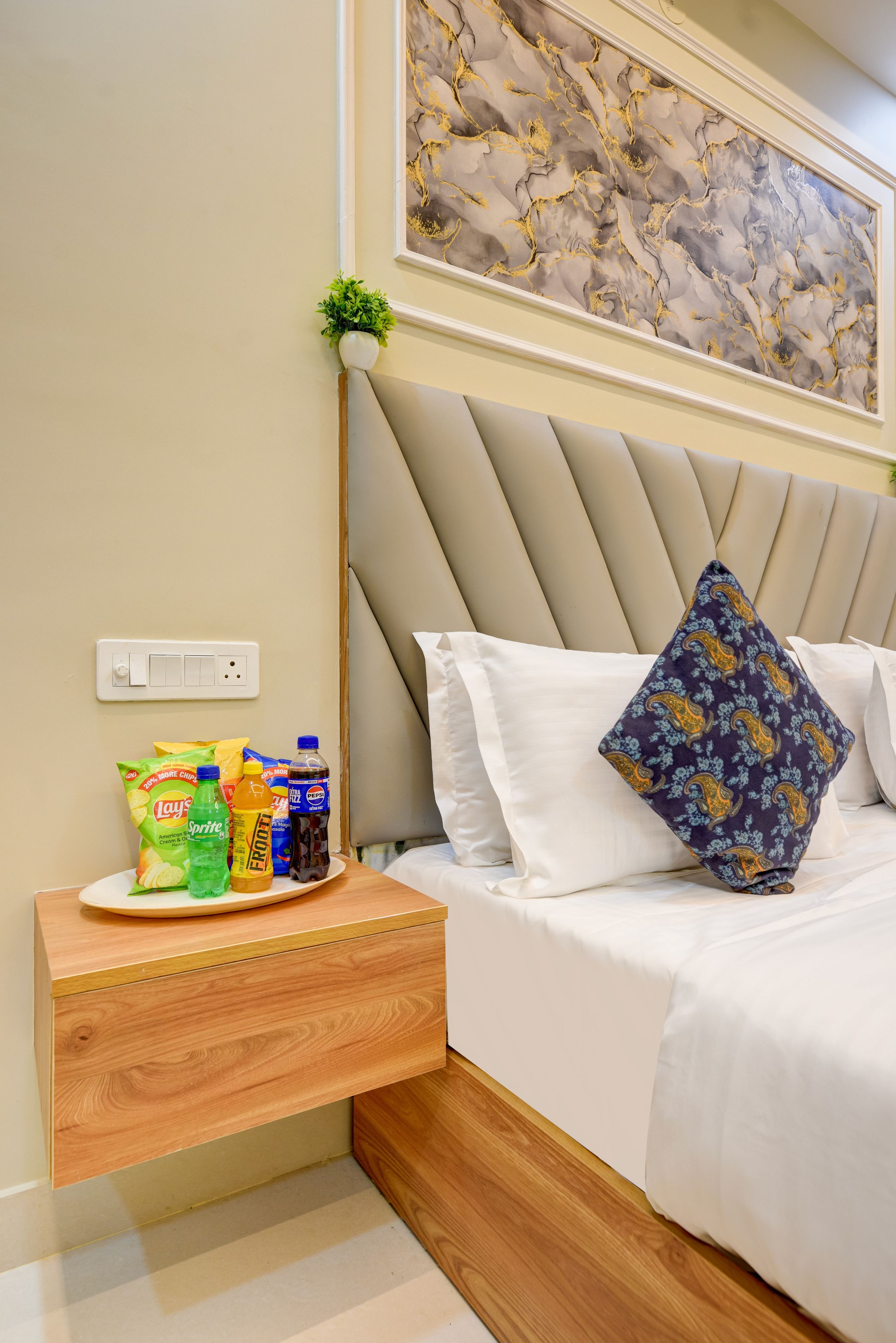 A hotel room with a bed, a bedside table with snacks, a power outlet, and a wall-mounted artwork.