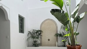 Um pátio com piso de azulejos, uma grande planta em vaso, uma mesa de madeira com uma bandeja de copos e um sofá verde.