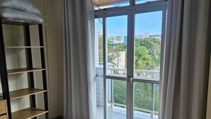 A bedroom with a bed, a shelf unit, and a view of a balcony and greenery. - Cabo Frio Seasonal Apartment - 3 minutes from the beach. Ideal for families (Vila Nova Cabo Frio)