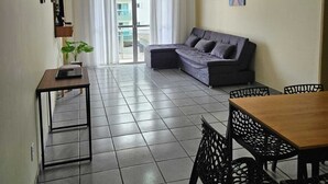A living room with a sofa, a dining table with chairs, a ceiling fan, and a window with curtains. - Cabo Frio Seasonal Apartment - 3 minutes from the beach. Ideal for families (Vila Nova Cabo Frio)