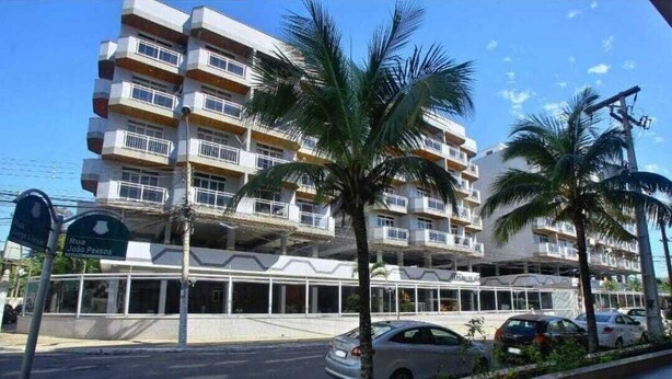 A multi-story hotel with balconies, palm trees, and a parking area. - Cabo Frio Seasonal Apartment - 3 minutes from the beach. Ideal for families (Vila Nova Cabo Frio)