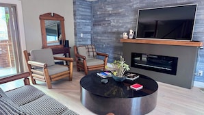 Uma sala de estar com lareira, dois poltronas xadrez, uma mesa de centro redonda e uma televisão de tela plana grande.