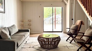 Uma sala de estar moderna com um sofá, uma mesa de centro redonda e uma cadeira. Há uma janela grande com vista para árvores e pedras.