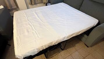 Una cama pequeña y compacta con colchón blanco y estructura metálica, ubicada entre dos sillones grises en una habitación con suelo de baldosas.
